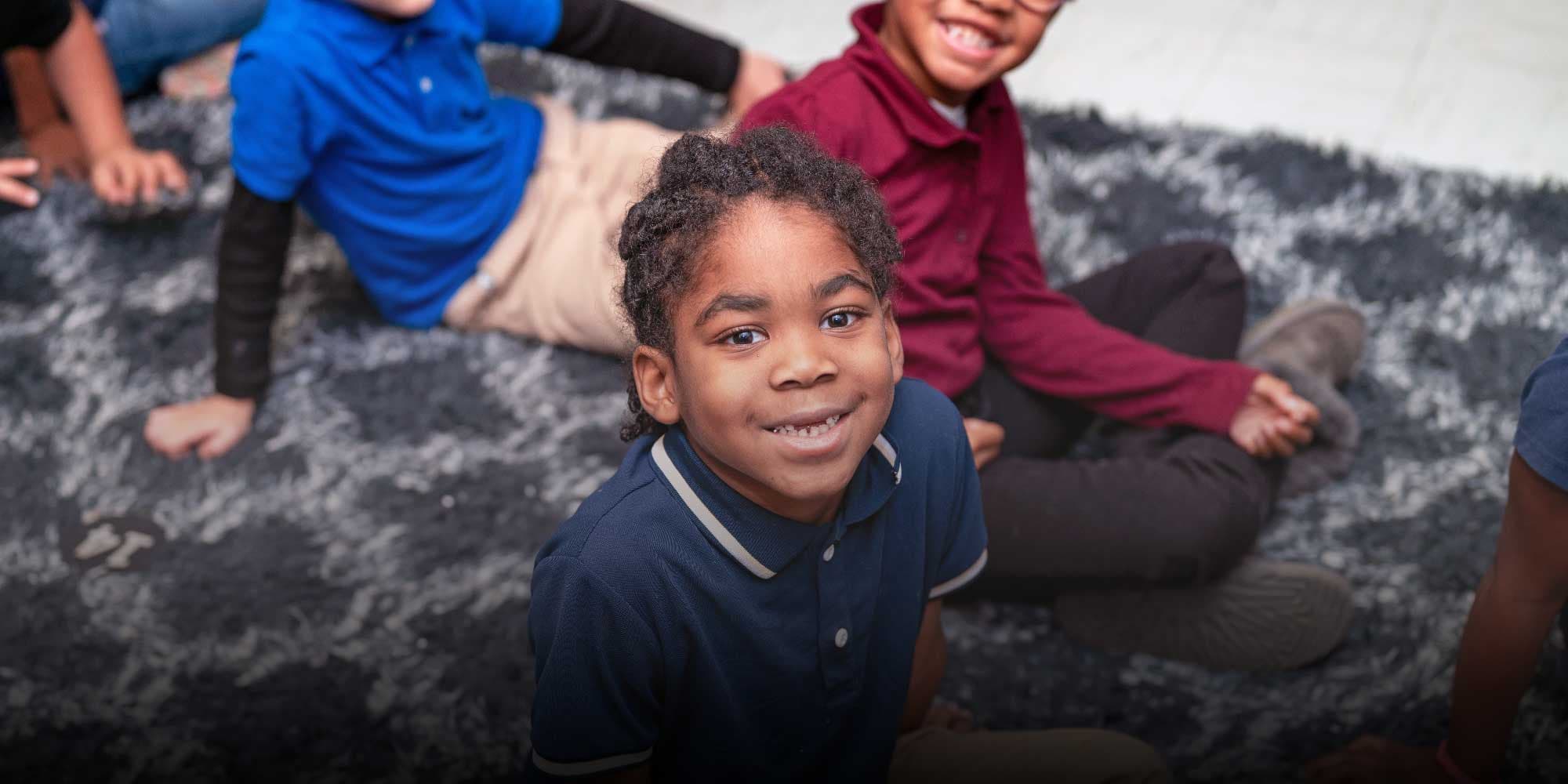 Smiling student in a blue polo sitting on a rug with classmates.